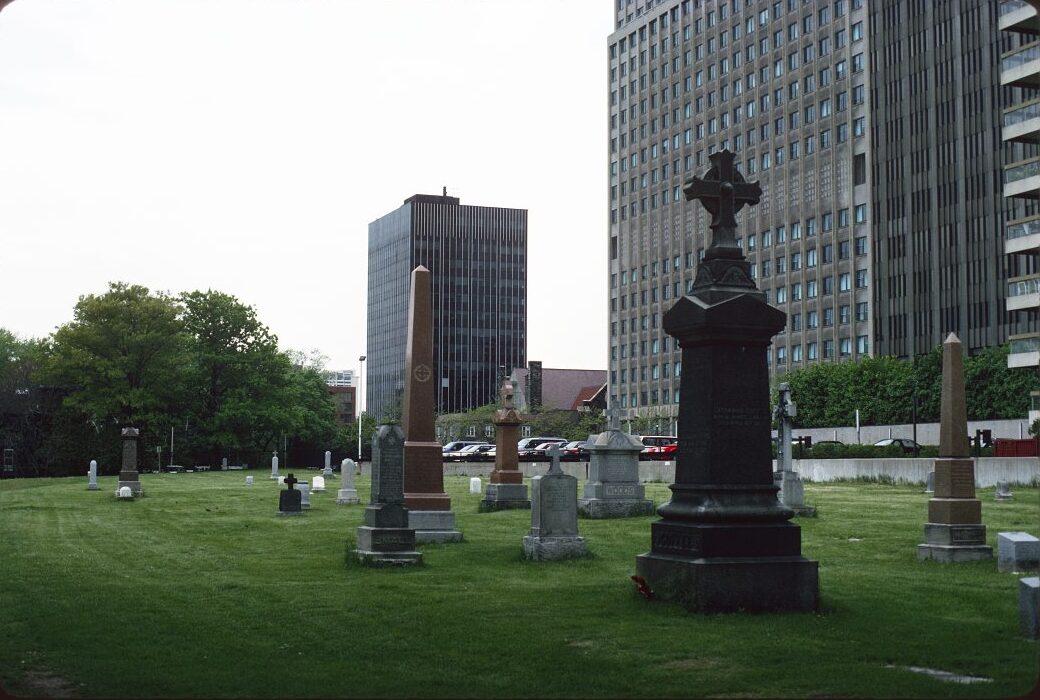 A cemetery with gravestones on a neat green lawn. In the background are several high-rise buildings.