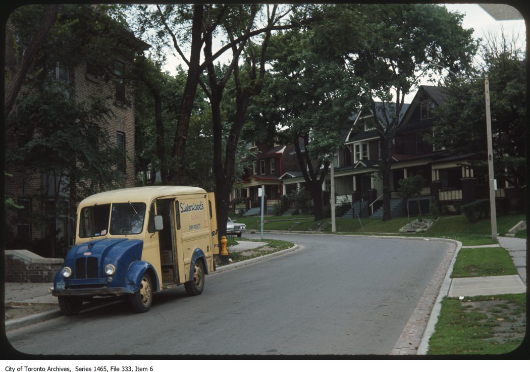 Blue and yellow van parked on a street with drive side door open. Street starts at foreground of image and carries through turning to the left. Homes lines the street.