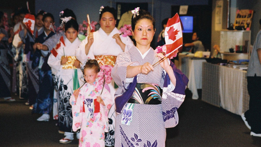 A group of women wearing walking in a line wearing traditional outfits holding Canadian flags.