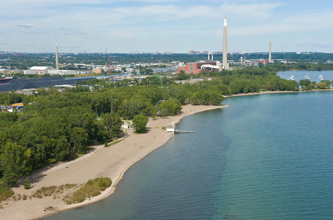 Aerial view of a beach and green spaces. Industrial chimneys and buildings are prominent in the background.