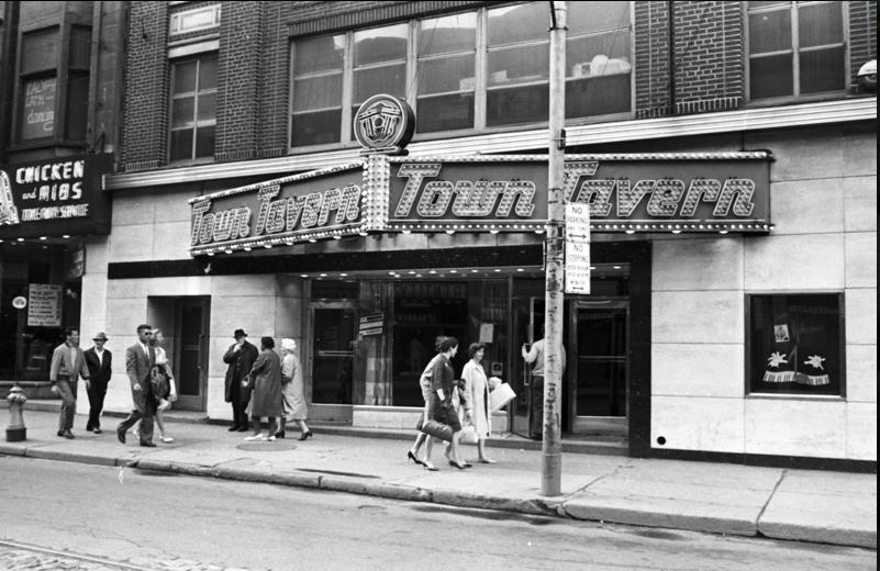 A black and white photograph of the Town Tavern's sign taken during the day with people walking by.