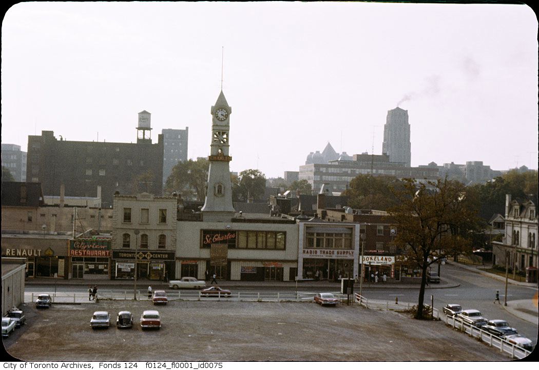 An urban vista with a large empty lot in the foreground, behind which stands a series of low-rise buildings. In the center, rising above building that features a red sign stating "St. Charles", is a tall white clock tower.