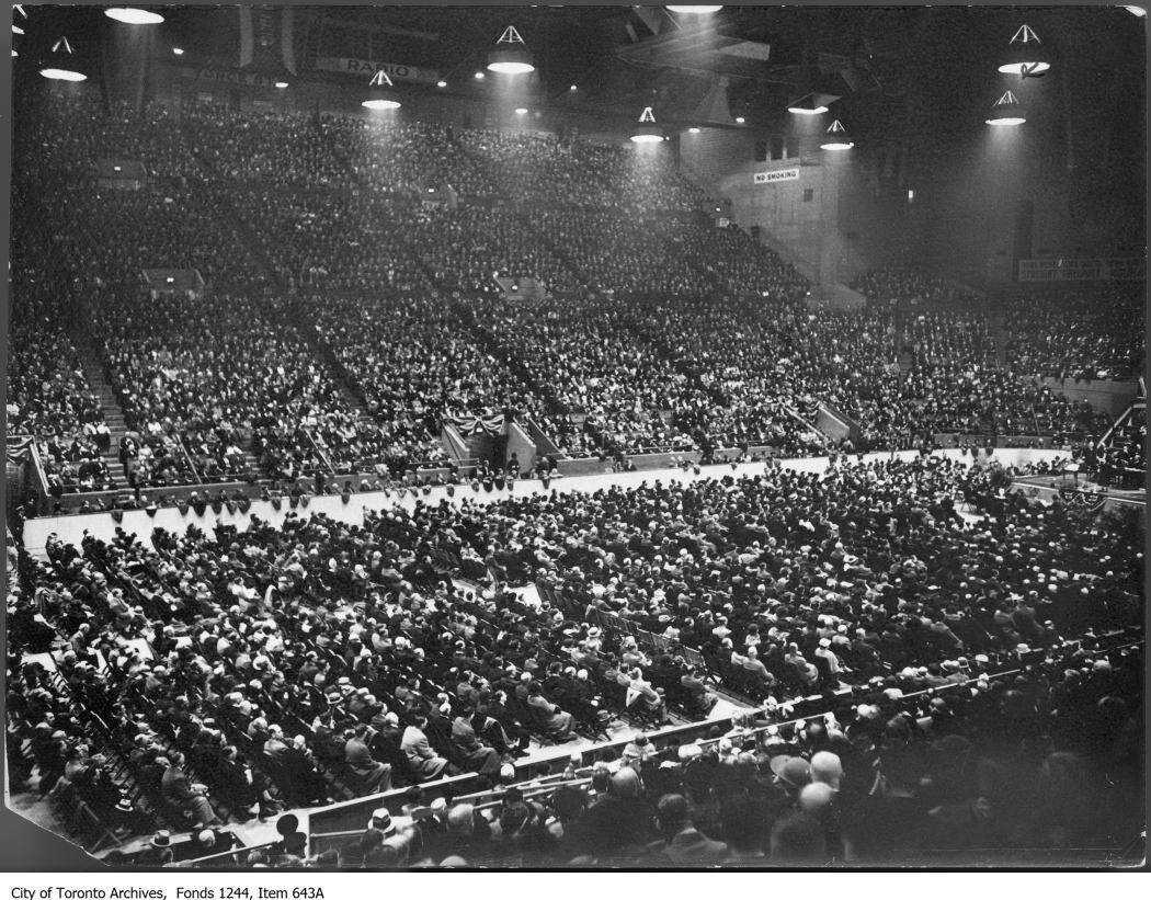 A black and white photograph of a large crowd seated in stands and in rows of neatly arranged chairs.  The audience is facing a small stage that is just barely visible at the right-edge of the photograph.