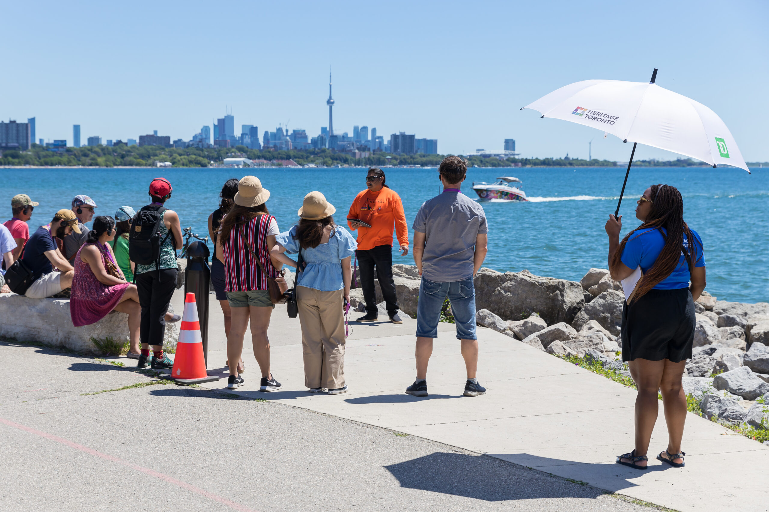 Group of people stand and sit along a path that fronts a waterfront view with a city skyline showing the CN tower behind. A person carrying a white umbrella appears on the right.