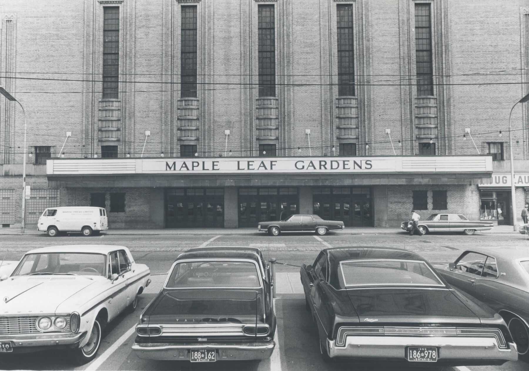 Exterior of an imposing building with a maruqee that reads "Maple Leaf Gardens". In the foreground are four parked cars.