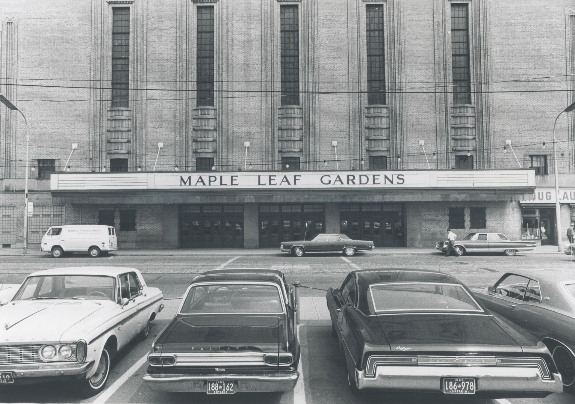 Exterior of an imposing building with a maruqee that reads "Maple Leaf Gardens". In the foreground are four parked cars.