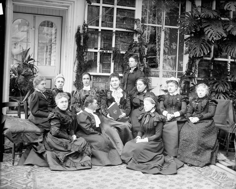 Eleven women pose for a formal black-and-white photograph. They are arranged in three rows: with three women in the foreground seated on the ground; seven women seated on chairs behind them; one woman stands at the back in the centre. All are wearing dark clothing, including long-sleeved blouses and long skirts. They are indoors, likely in a residential area.